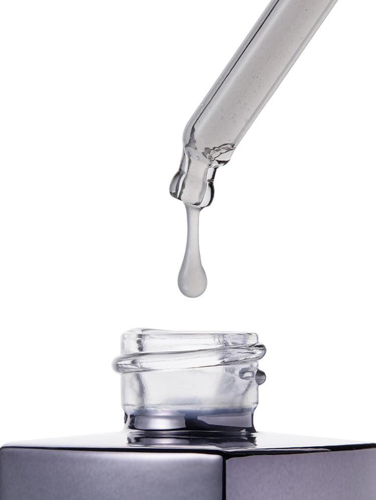 A clear glass dropper tube dispensing a single transparent liquid drop above an open clear glass bottle with a dark metallic cap, isolated on a white background.