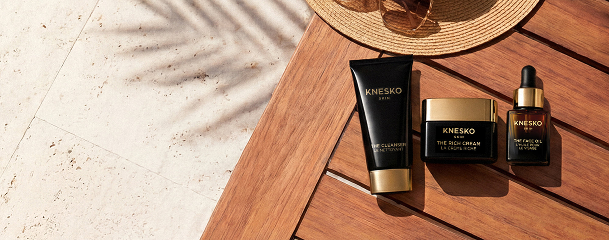 Three Knesko skincare products arranged on a wooden table outdoors: a black tube with a gold base, a black jar with a gold lid, and a small black dropper bottle with a gold cap. A straw hat and sunglasses sit behind them, with palm leaf shadows cast across the scene.
