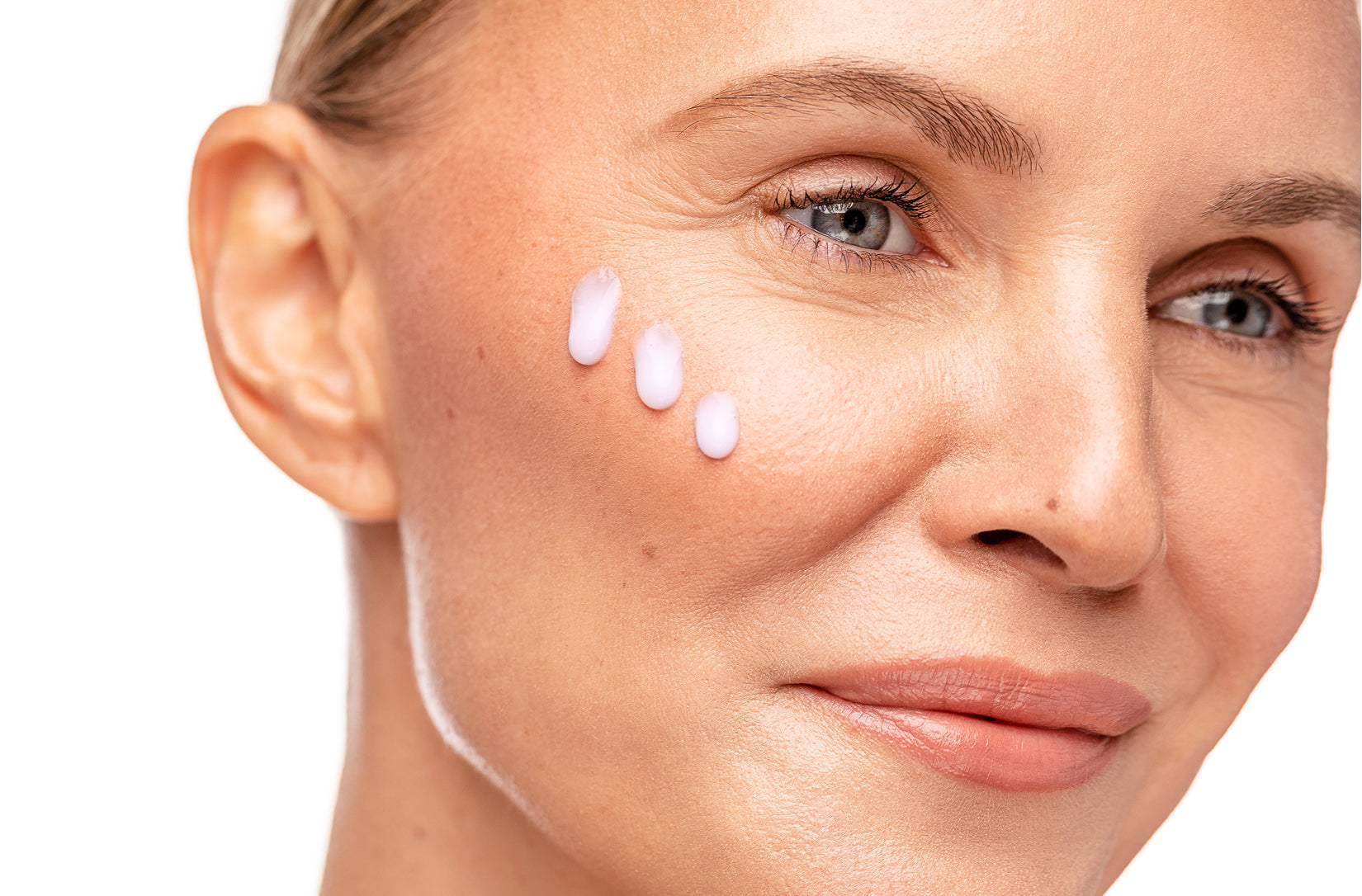 Close-up of a woman's face with three small drops of product applied to her cheek, shown against a clean white background.