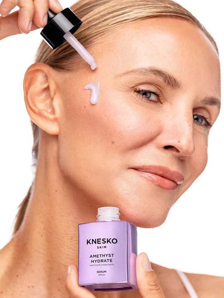 A close-up of a woman applying a white serum from a black dropper onto her cheek, with the Knesko Skin Amethyst Hydrate serum bottle held in her other hand against a white background.