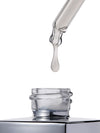 A clear glass dropper with a single drop of transparent liquid hanging above an open clear bottle on a white background.