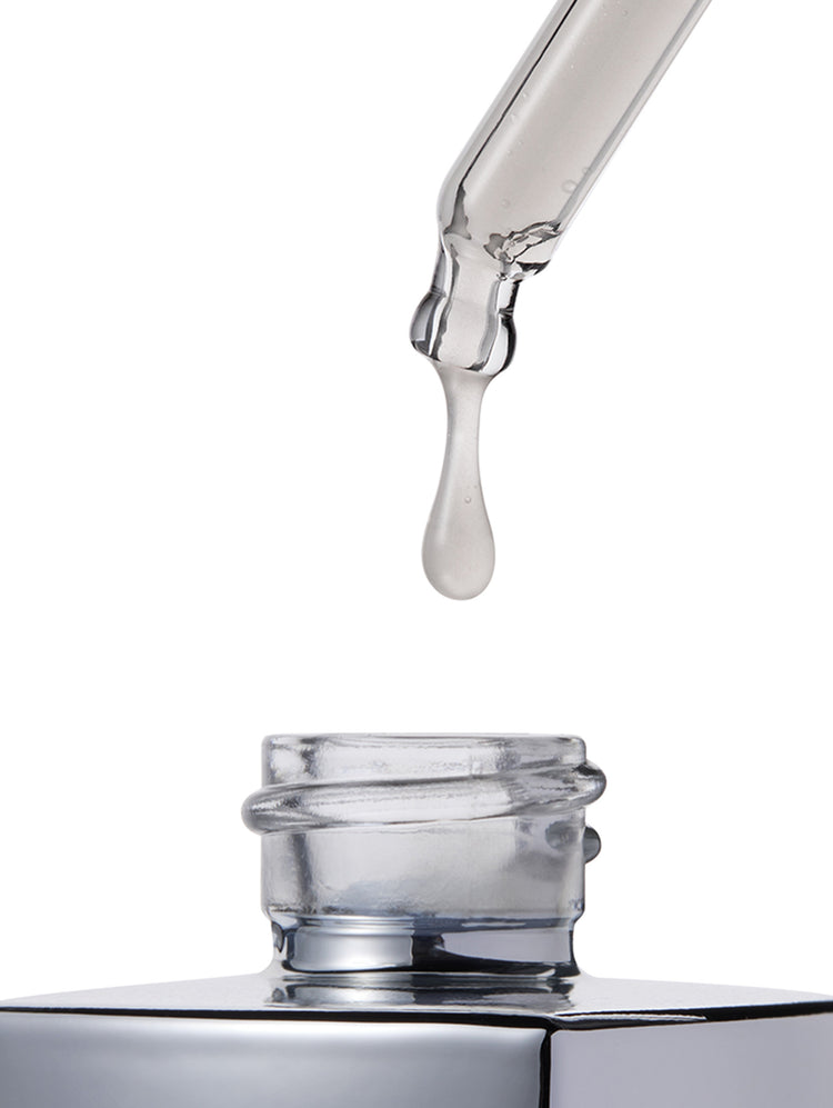 A clear glass dropper with a single drop of transparent liquid hanging above an open clear bottle on a white background.