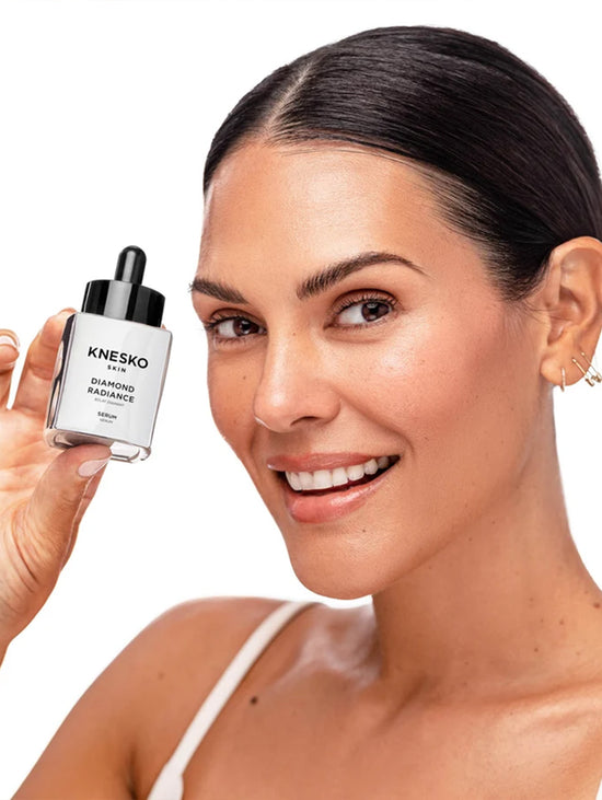 A smiling woman with dark hair pulled back holds up a small Knesko skincare dropper bottle against a clean white background.