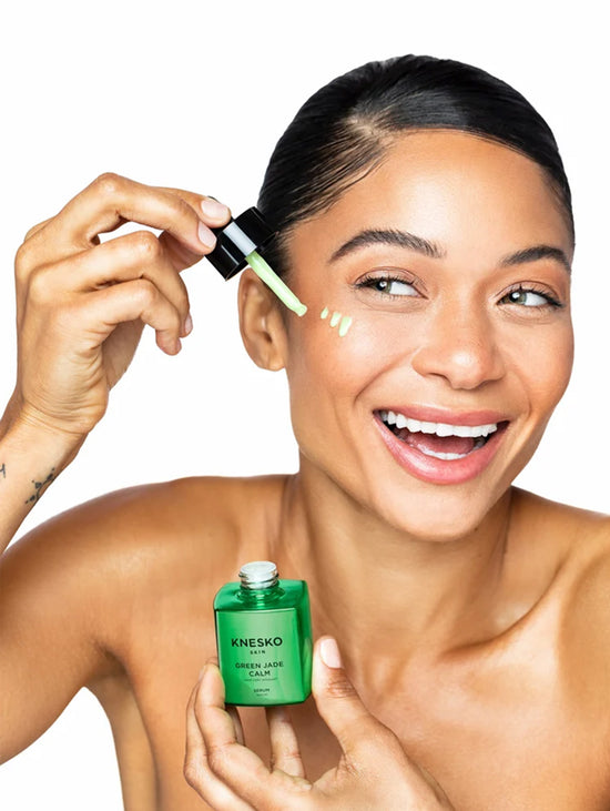 A smiling woman with dark hair pulled back applies a green cream from a dropper to her cheek while holding a green Knesko serum bottle against a white background.