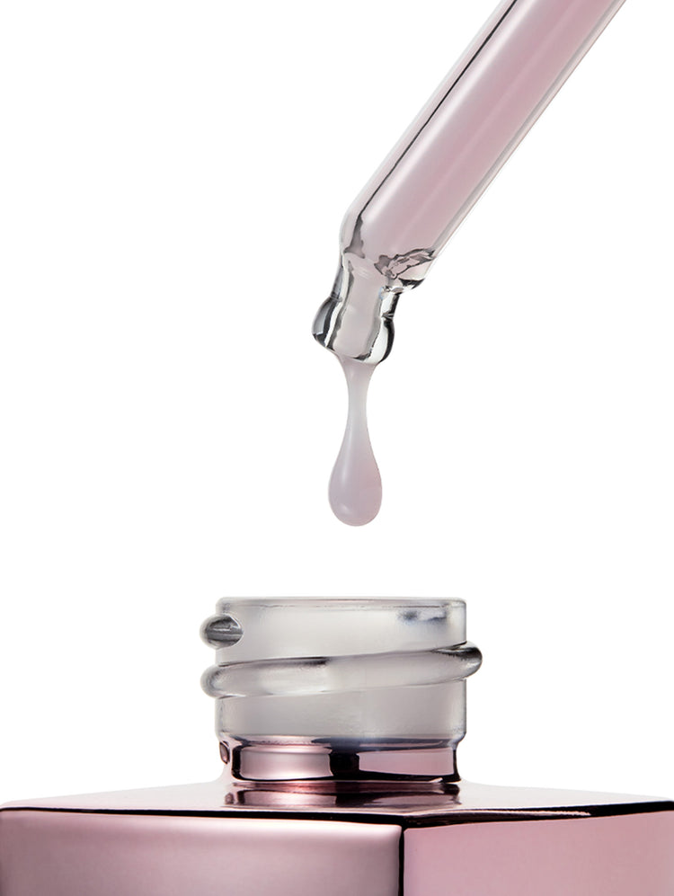 A close-up of a clear glass dropper above an open pink cosmetic bottle, with a single pale pink liquid drop hanging in the center against a white background.