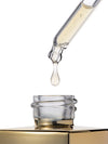 A clear glass dropper holding a drop of pale golden liquid above an open gold-toned bottle cap on a white background.