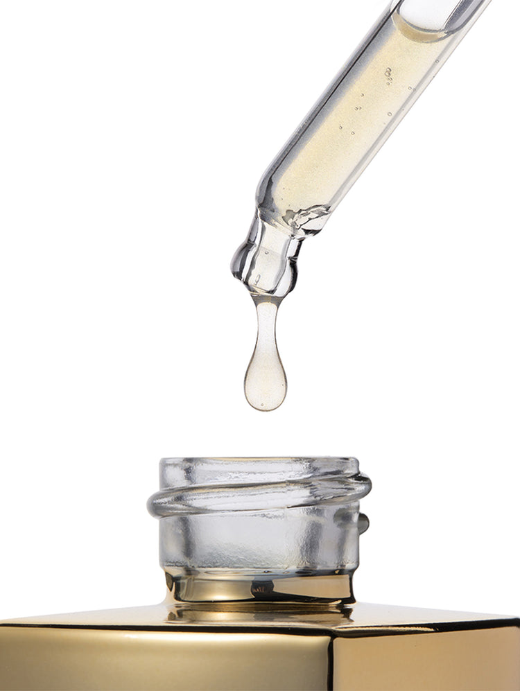 A clear glass dropper holding a drop of pale golden liquid above an open gold-toned bottle cap on a white background.