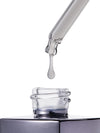 A clear glass dropper tube dispensing a single transparent liquid drop above an open clear glass bottle with a dark metallic cap, isolated on a white background.