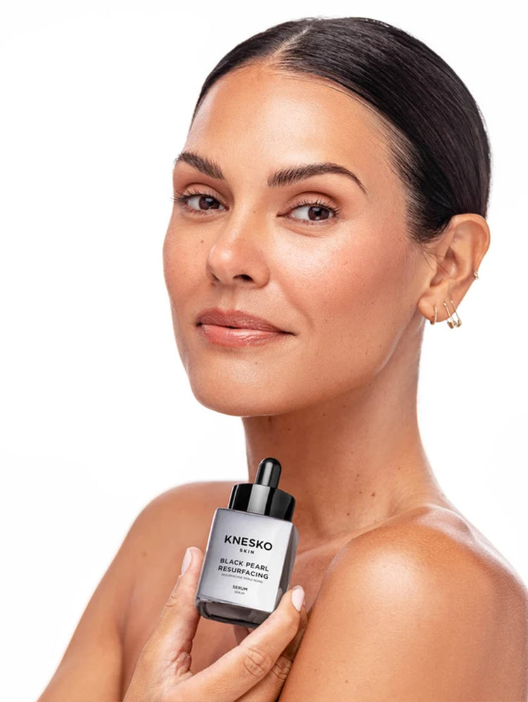 A woman with dark hair pulled back holds a bottle of KNESKO Pearl Resurfacing Serum against a clean white background.