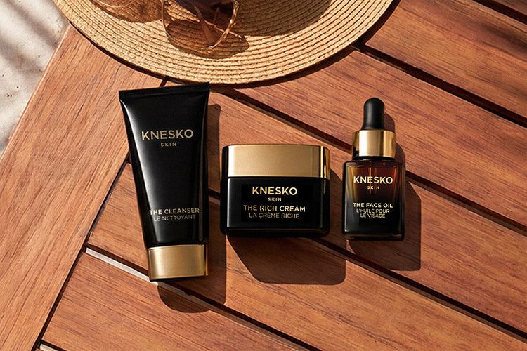 Three Knesko Skin skincare products arranged on a wooden surface: a black cleanser tube, a black rich cream jar with a gold lid, and a black face oil bottle with a gold dropper cap. A straw hat is partially visible at the top.
