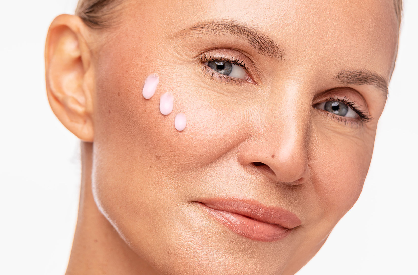 Close-up of a woman's face with three small dollops of product applied to her cheek, set against a clean white background.