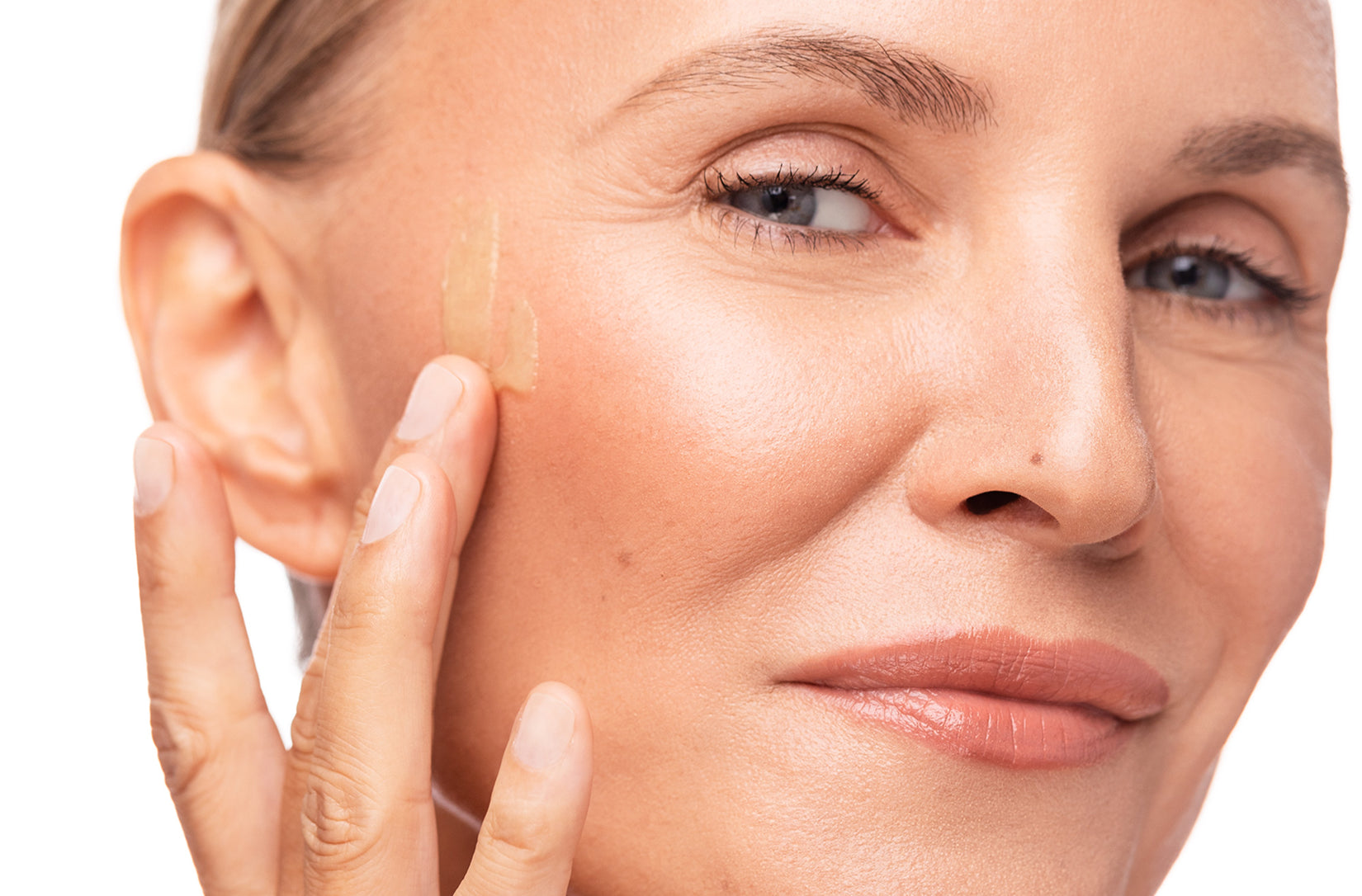 Close-up of a woman applying a small amount of translucent product to her cheek against a clean white background.